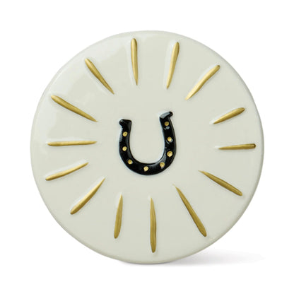 A round off-white decorative plaque or coaster featuring a black horseshoe in the center with small gold dots, surrounded by radiating gold lines on a white background.