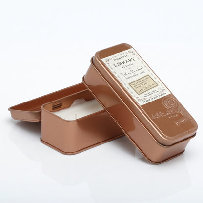 A rectangular metal tin candle shown open with the lid leaning against it on a clean white background. The tin has a warm metallic copper/rose-gold finish and a vintage-style label on the lid reading &