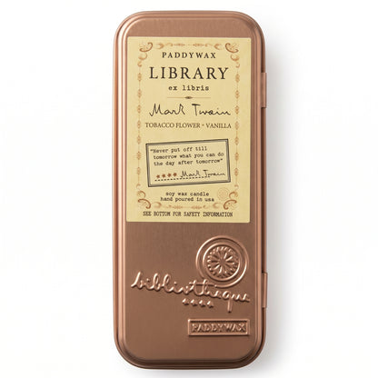 A rectangular metal tin of Paddywax Library ex Libris top wax candle, featuring a warm copper-bronze finish and a cream-colored label with black text. The label details the scent as &