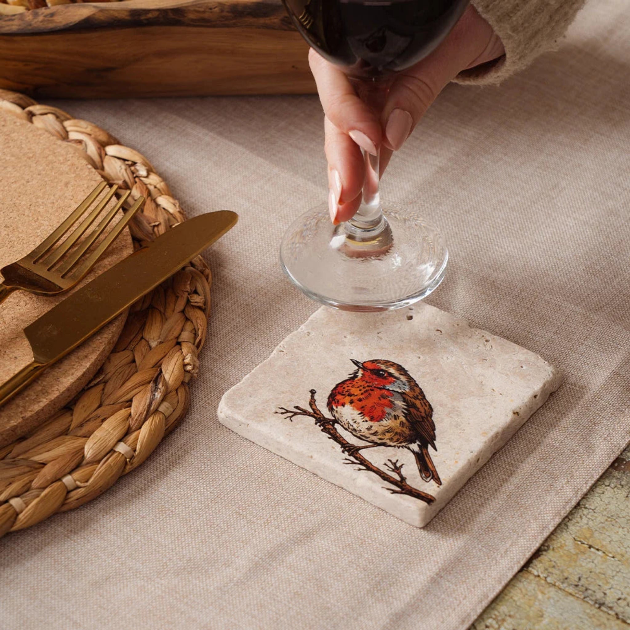 Hand placing a glass on a coaster with a robin design, next to a woven basket and gold knife.