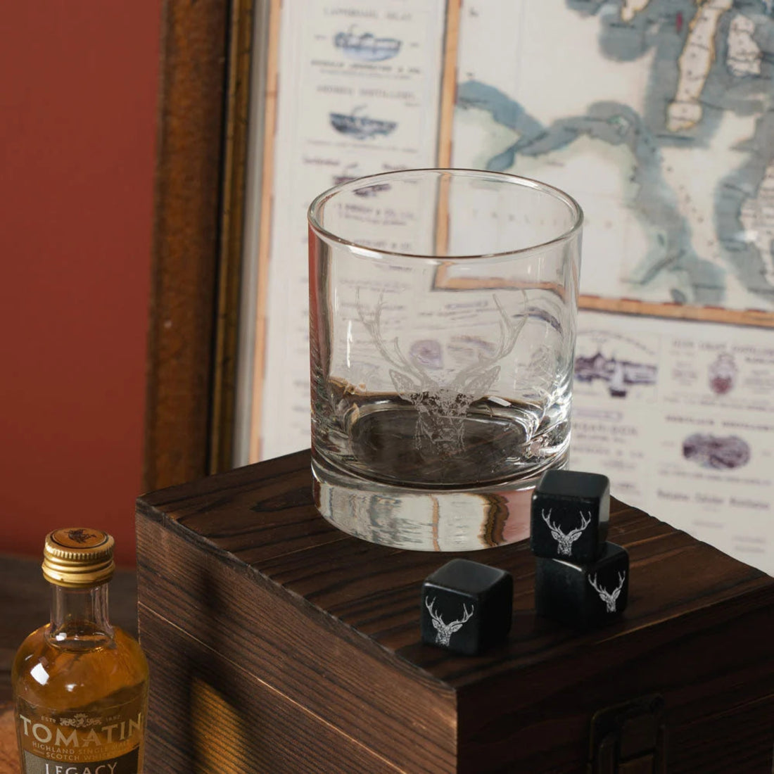 Clear glass with Stag engraving, black stones, and a bottle of Tomatin Legacy whisky on a wooden surface with a map in the background.