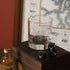 Clear glass with Stag engraving, black stones, and a bottle of Tomatin Legacy whisky on a wooden surface with a map in the background.