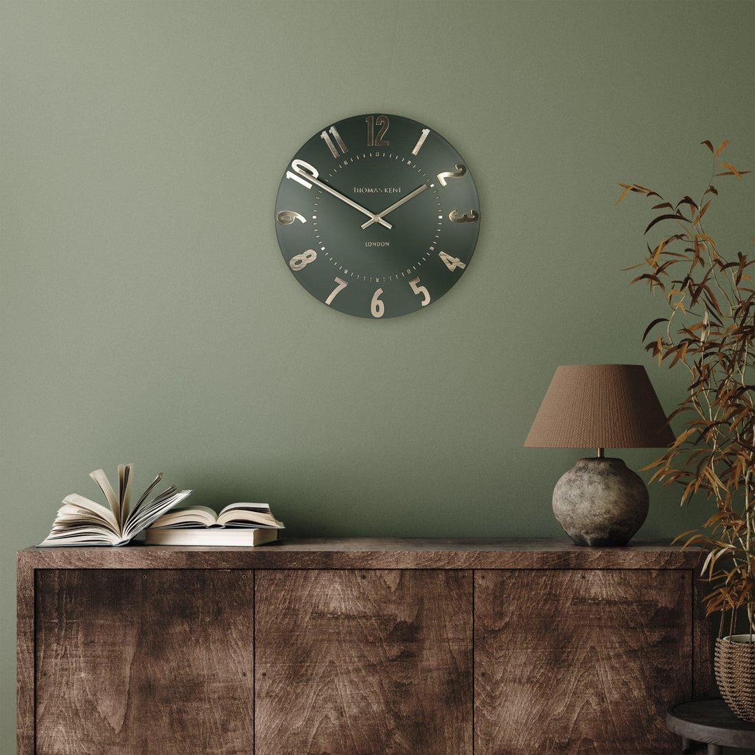 Wall clock on a green wall above a wooden cabinet with books and a lamp.