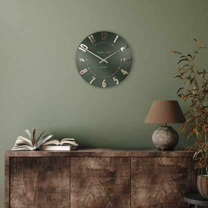 Wall clock on a green wall above a wooden cabinet with books and a lamp.