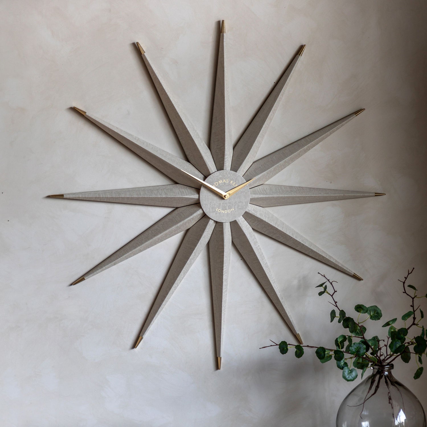 Decorative wall clock with starburst design on a light wall, accompanied by a vase with greenery.