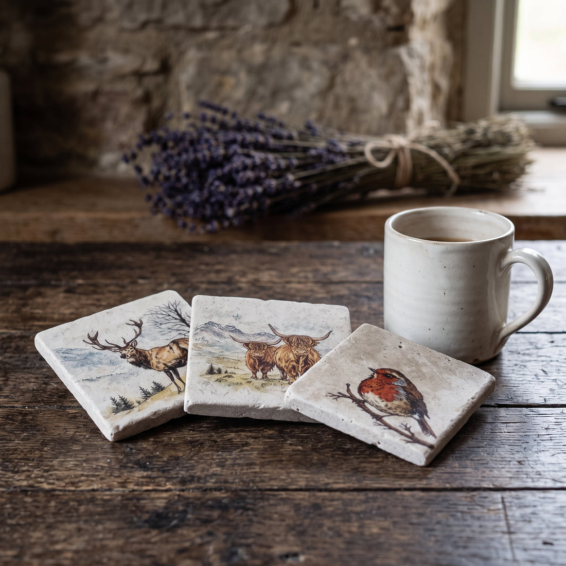Three square ceramic coasters with nature illustrations (stag, highland cow, robin) are arranged on a rustic wooden table next to a white ceramic mug, with a blurred background of a stone wall and lavender.