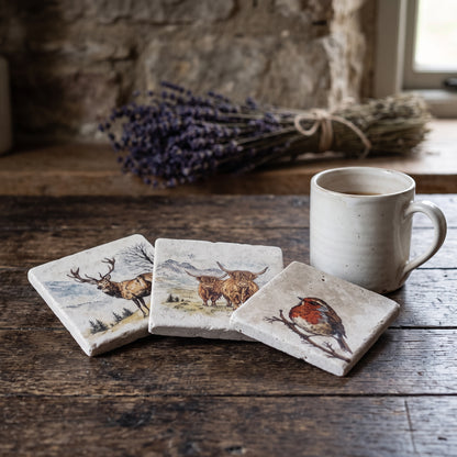 Three square ceramic coasters with nature illustrations (stag, highland cow, robin) are arranged on a rustic wooden table next to a white ceramic mug, with a blurred background of a stone wall and lavender.