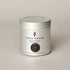A silver metal tin candle with a matching lid, featuring a white label with black text and a circular amber-colored scent badge. The brand &