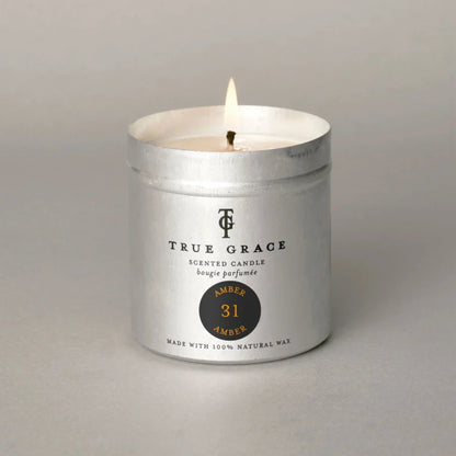 A lit True Grace scented candle in a silver metal tin with a white wax fill, set against a neutral gray background.