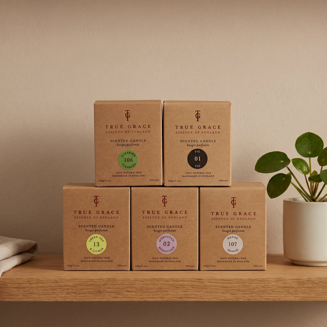 Five True Grace scented candles in brown kraft paper boxes are arranged on a wooden shelf against a beige wall, with a small potted plant on the right and a folded cloth on the left.