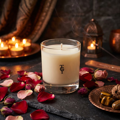 A white wax candle in a clear glass jar sits on a dark stone surface surrounded by red rose petals, dried roses, and small lit candles in the background. A black monogram logo is printed on the front of the jar.