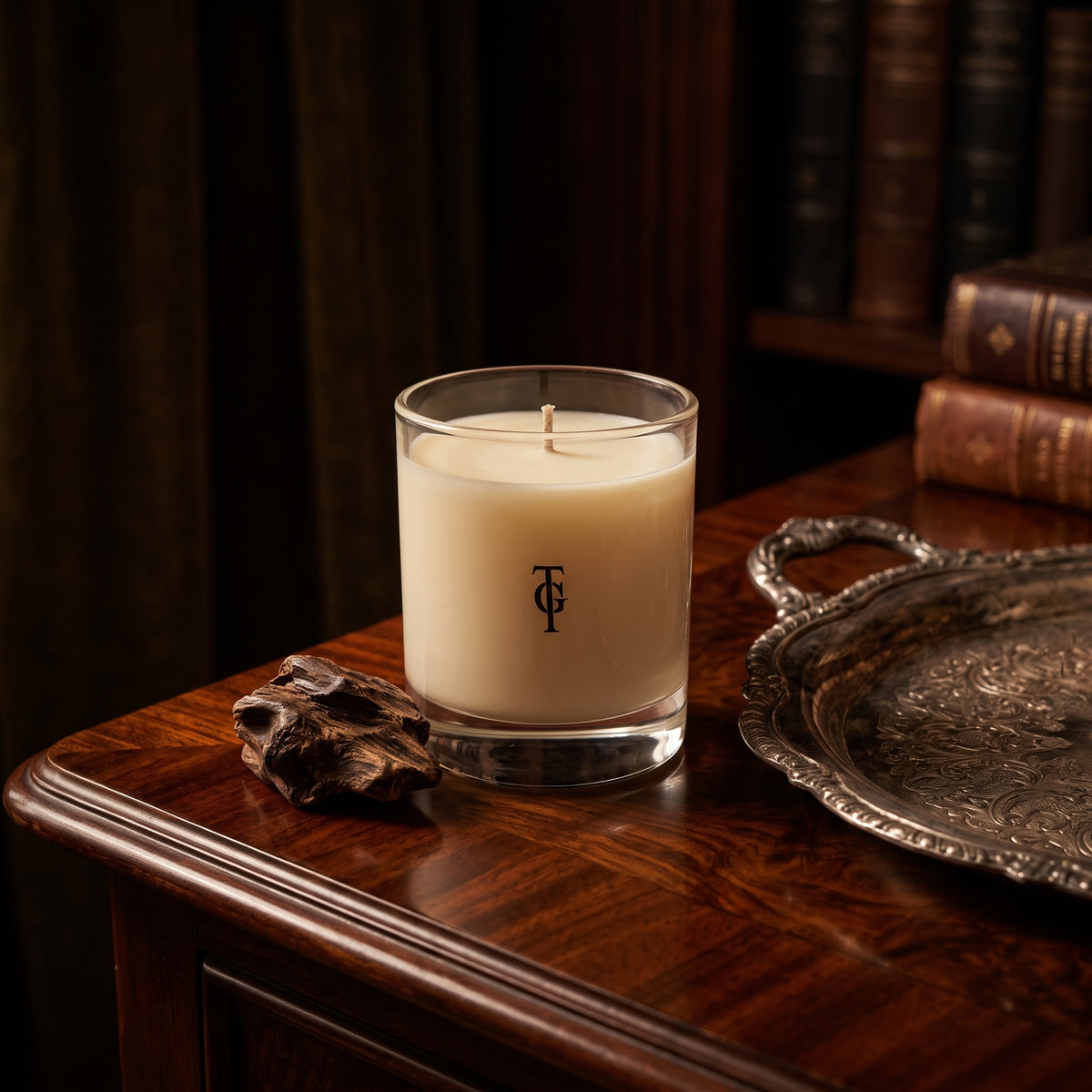A cream-colored scented candle in a clear glass jar with a black monogram logo, set on a dark wooden table next to a silver tray and a piece of driftwood, with a blurred background of books.
