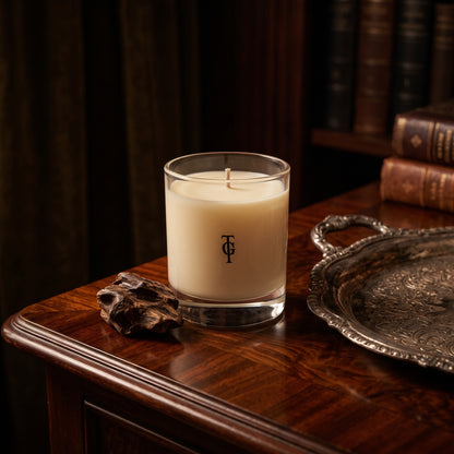 A cream-colored scented candle in a clear glass jar with a black monogram logo, set on a dark wooden table next to a silver tray and a piece of driftwood, with a blurred background of books.