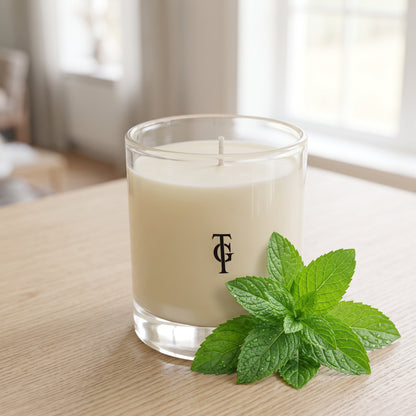 A clear glass jar candle with creamy off-white wax sits on a light wood table, with a sprig of fresh green mint leaves placed in front. The jar has a black monogram logo on the front.