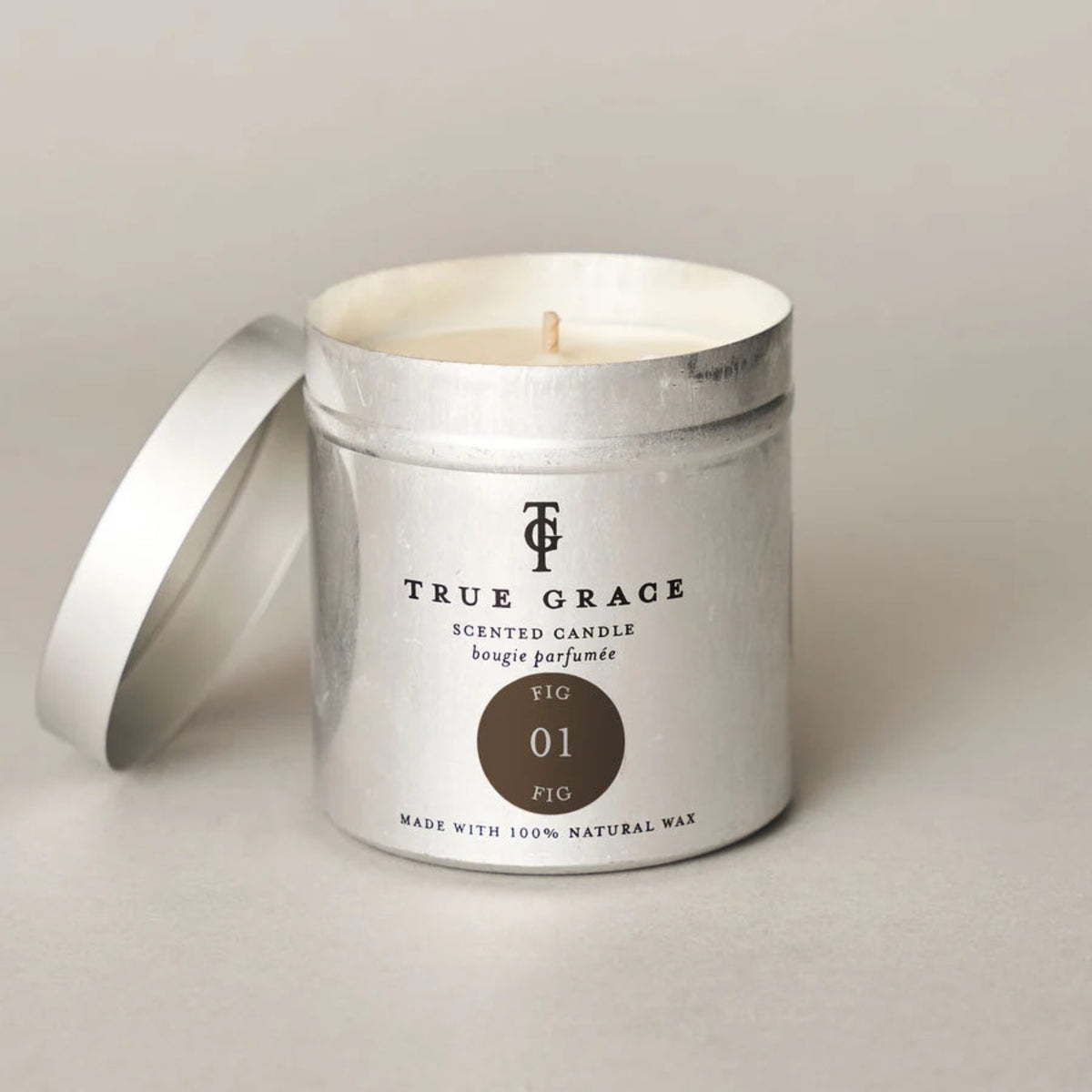 A silver metal scented candle tin with its lid leaning beside it on a light neutral background. The front label reads TRUE GRACE and shows a circular brown shade callout labeled FIG 01.
