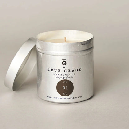 A silver metal scented candle tin with its lid leaning beside it on a light neutral background. The front label reads TRUE GRACE and shows a circular brown shade callout labeled FIG 01.