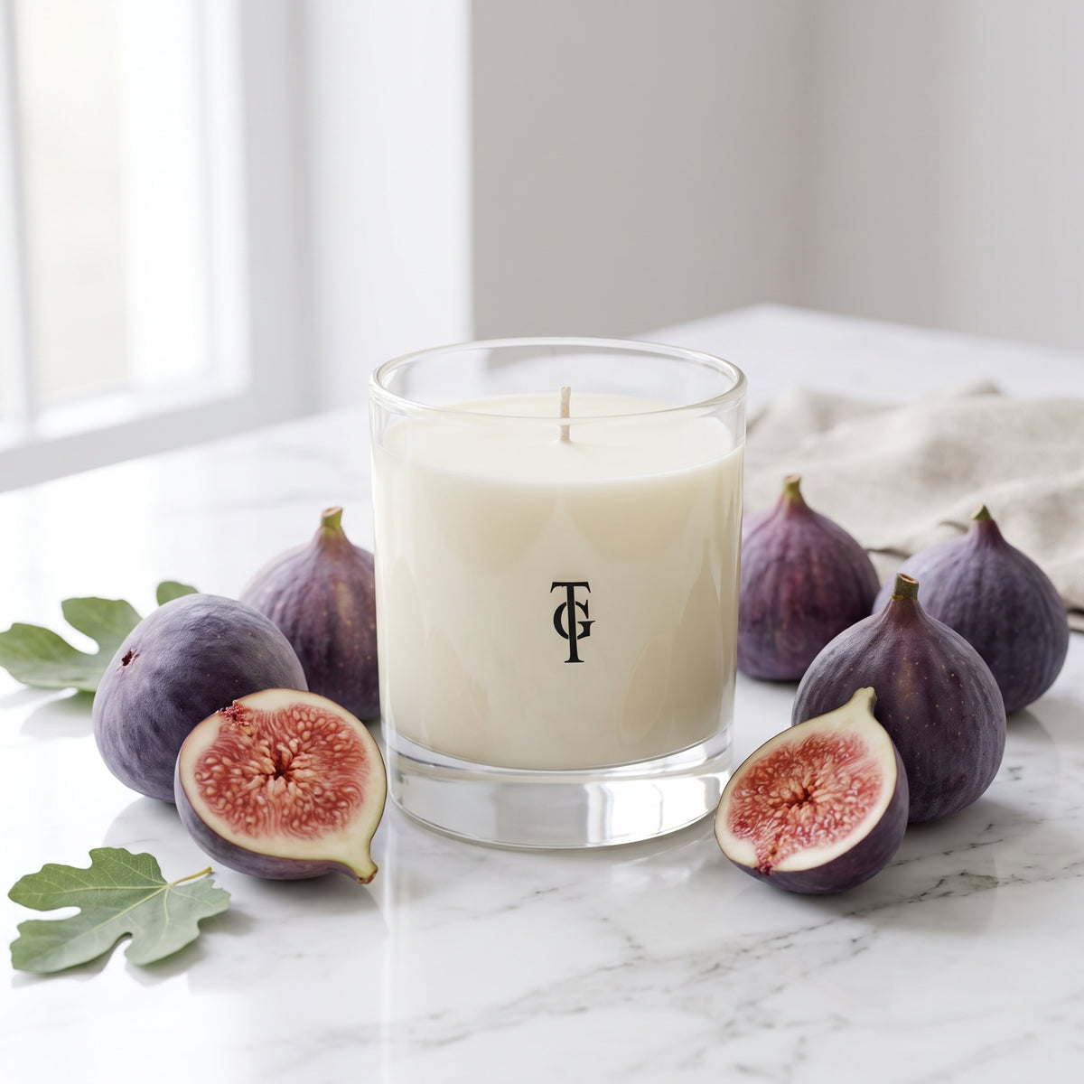 A white wax candle in a clear glass jar with a black monogram logo, styled on a white marble surface with fresh figs and green leaves.