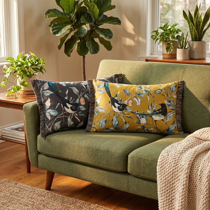 Two decorative cushions with bird and leaf patterns rest on an olive green sofa in a styled living room with potted plants and warm wood flooring.