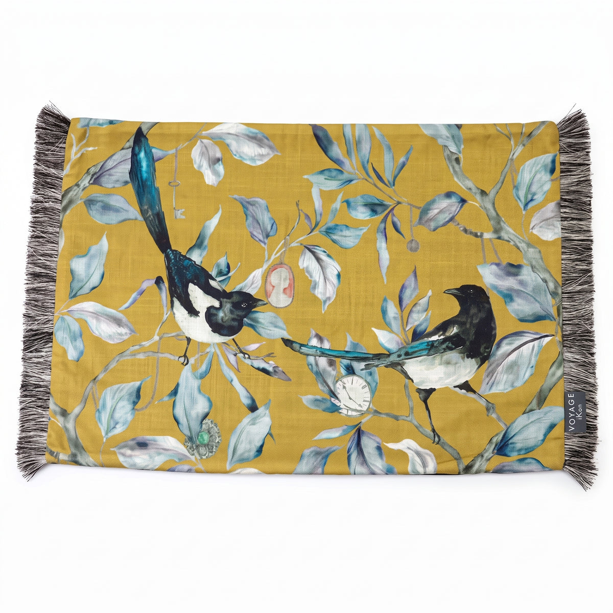 A rectangular woven textile with fringe on both short ends, featuring a mustard yellow background and an illustrated print of two magpies among blue-gray leaves and small birds. A small side tag reads &