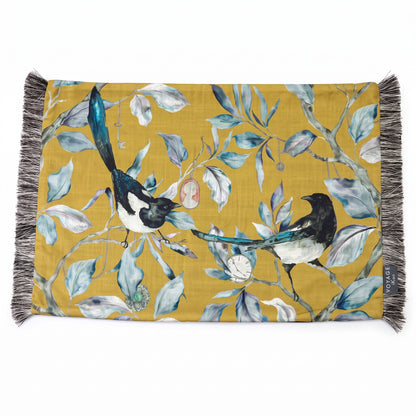 A rectangular woven textile with fringe on both short ends, featuring a mustard yellow background and an illustrated print of two magpies among blue-gray leaves and small birds. A small side tag reads &