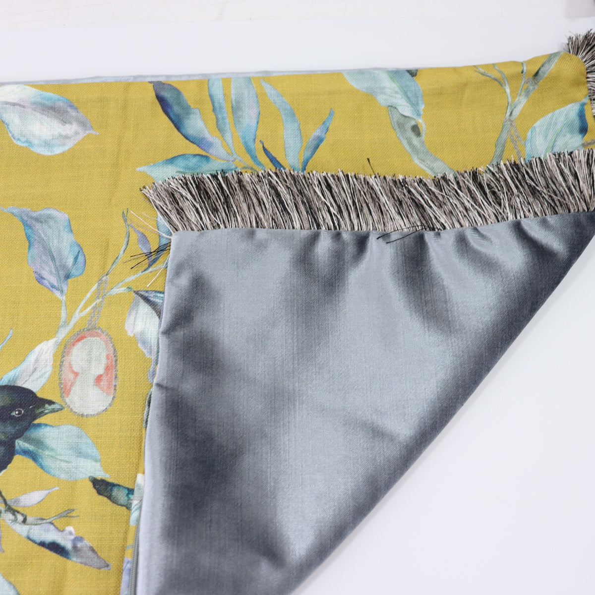 A close-up of a folded cushion with a mustard yellow botanical print featuring blue-green leaves, a small bird, and a pink fruit, with a gray satin reverse side and fringed edges.