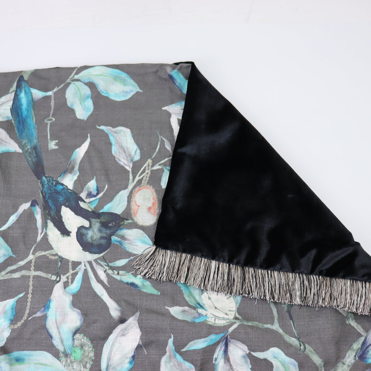 A folded cushion with a gray background featuring a watercolor-style bird and leaf pattern, with a black satin reverse side and fringe trim.