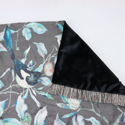 A folded cushion with a gray background featuring a watercolor-style bird and leaf pattern, with a black satin reverse side and fringe trim.