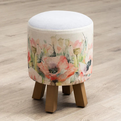 A round upholstered stool with a white cushion top and four wooden legs, wrapped in a floral fabric print featuring pink and peach poppies on a light beige background, set on a light wood floor.