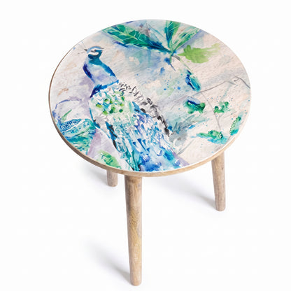 A round side table with a light natural wood frame and three legs, featuring a top printed with a vibrant watercolor painting of a peacock and green leaves on a light background.