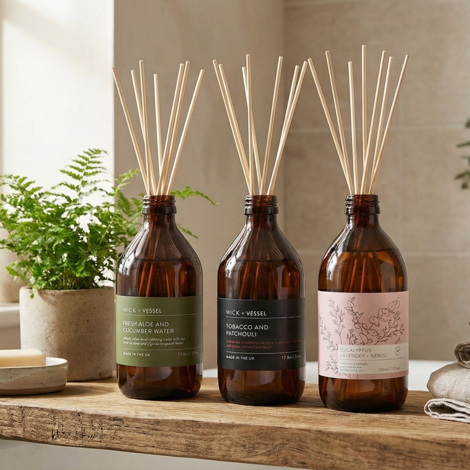 Three diffuser bottles on a wooden surface with plants and towels in the background