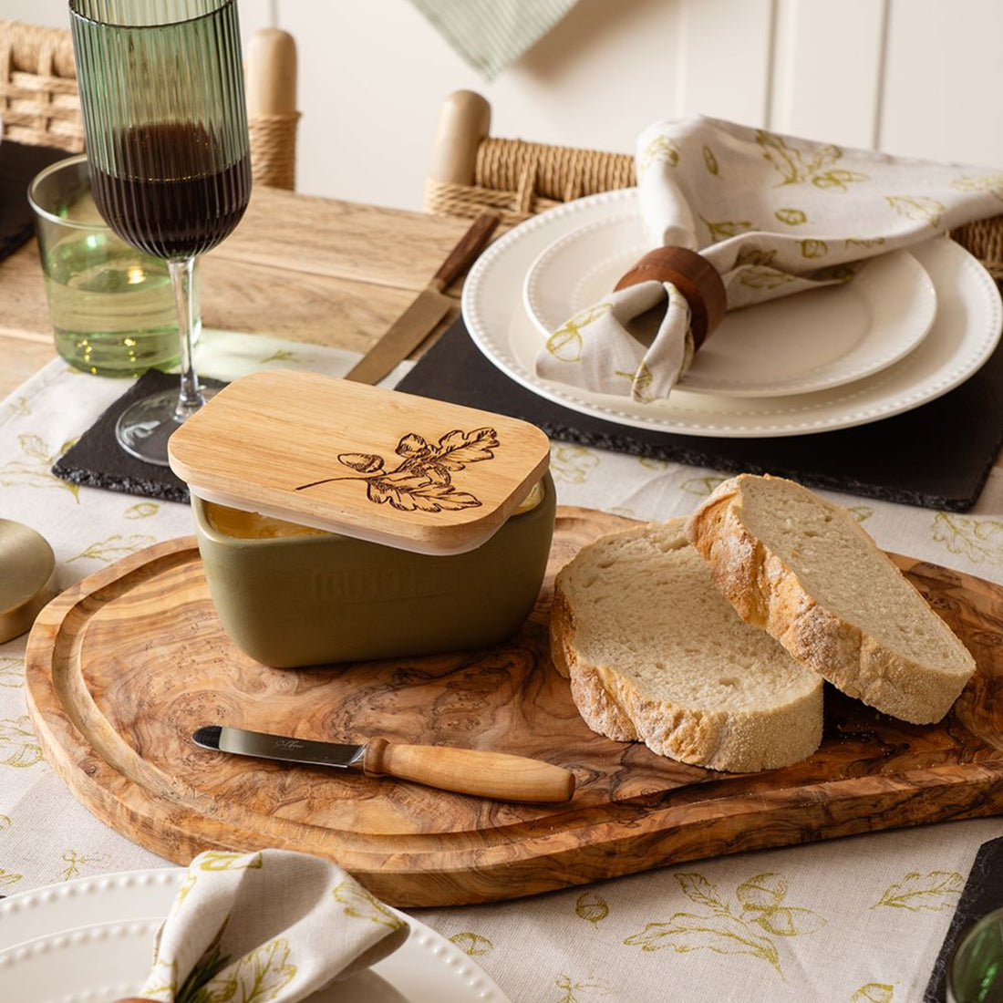 Butter dish with wooden lid on a wooden cutting board with bread slices, knife, and glasses of wine on a table.