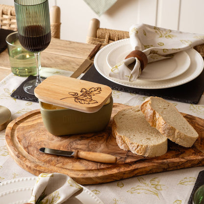 Butter dish with wooden lid on a wooden cutting board with bread slices, knife, and glasses of wine on a table.
