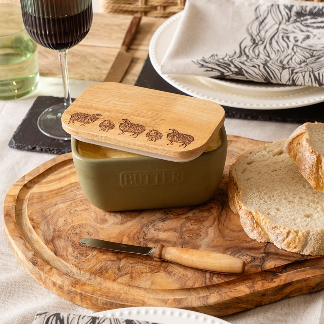 Butter container with wooden lid on a wooden cutting board with bread and a glass of red wine.