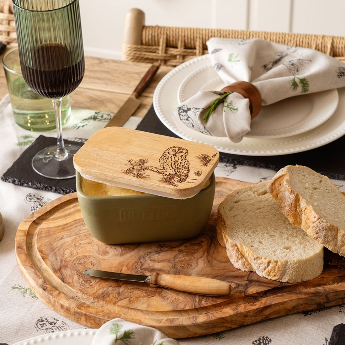 Green container with wooden lid on a wooden cutting board with bread slices, knife, and glasses of wine.