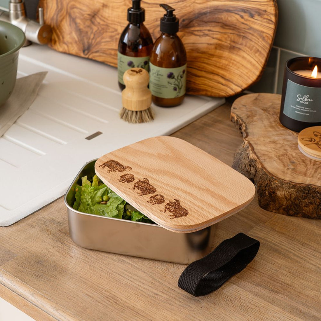 Metal lunch box with wooden lid containing salad, on a kitchen counter with cleaning supplies and a candle.