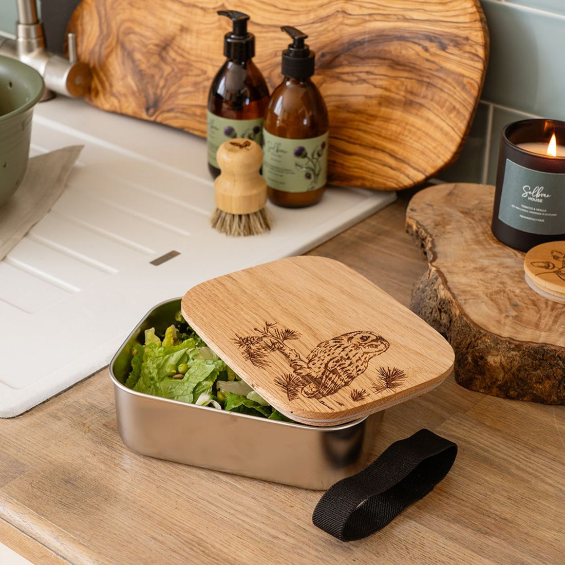 Metal lunch box with wooden lid containing salad, surrounded by natural products on a wooden surface.