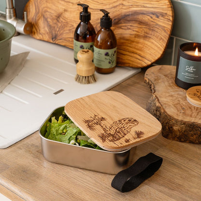 Metal lunch box with wooden lid containing salad, surrounded by natural products on a wooden surface.